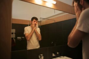 Man in white t-shirt reflects in bathroom mirror, appearing thoughtful and concerned.