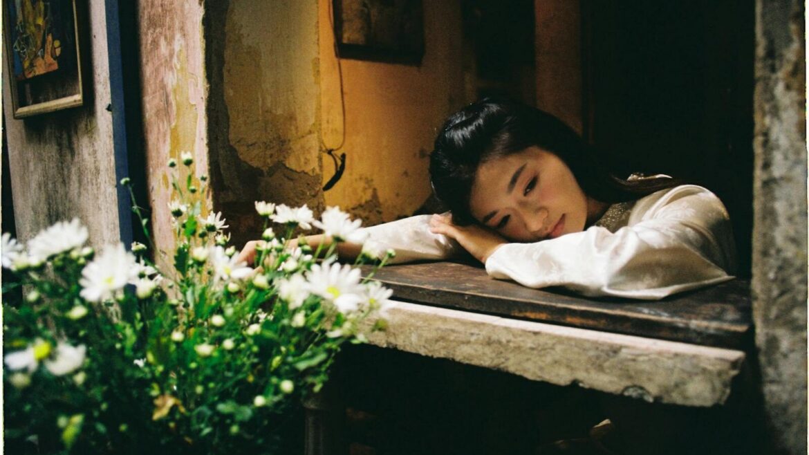 A contemplative woman rests by an open window with daisy flowers nearby.