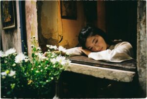 A contemplative woman rests by an open window with daisy flowers nearby.
